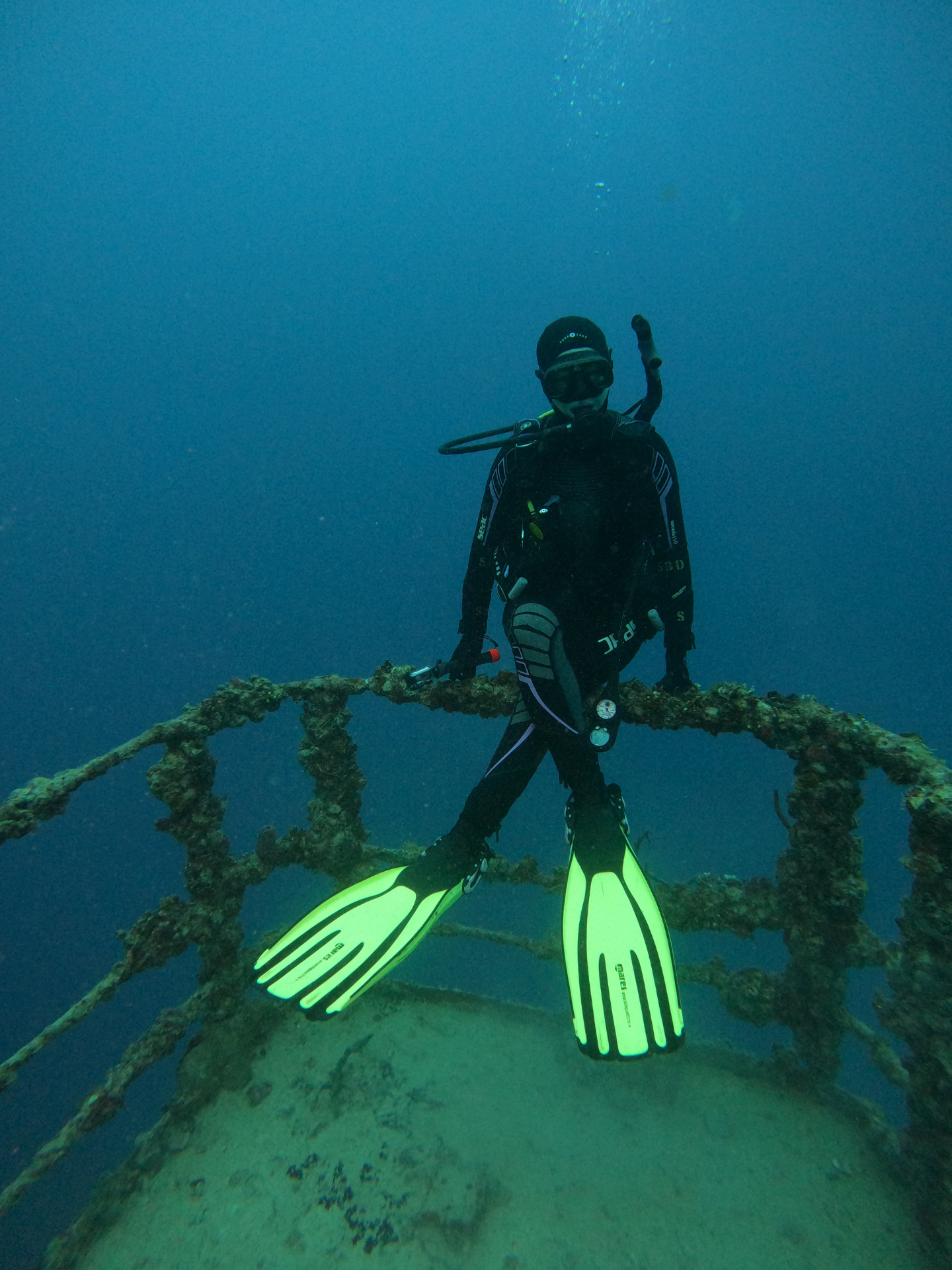 A scuba diver in a full black wetsuit, mask and tank sits with ankles crossed on the algae-covered railing of a sunken shipwreck, neon yellow-green fins bright against the deep blue water, bubbles rising toward the surface