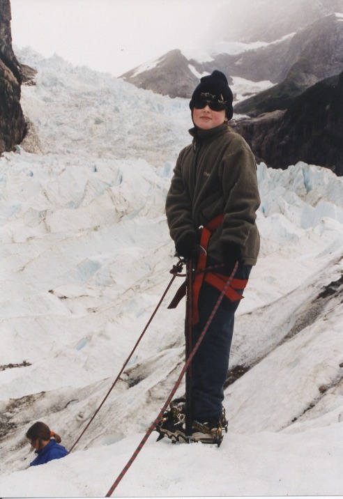 A child in a black beanie, sunglasses and olive-green fleece stands on a glacier with crampons on her boots and a red harness at her hips, both gloved hands resting on an ice axe planted in the snow, blue-white ice walls and misty peaks behind