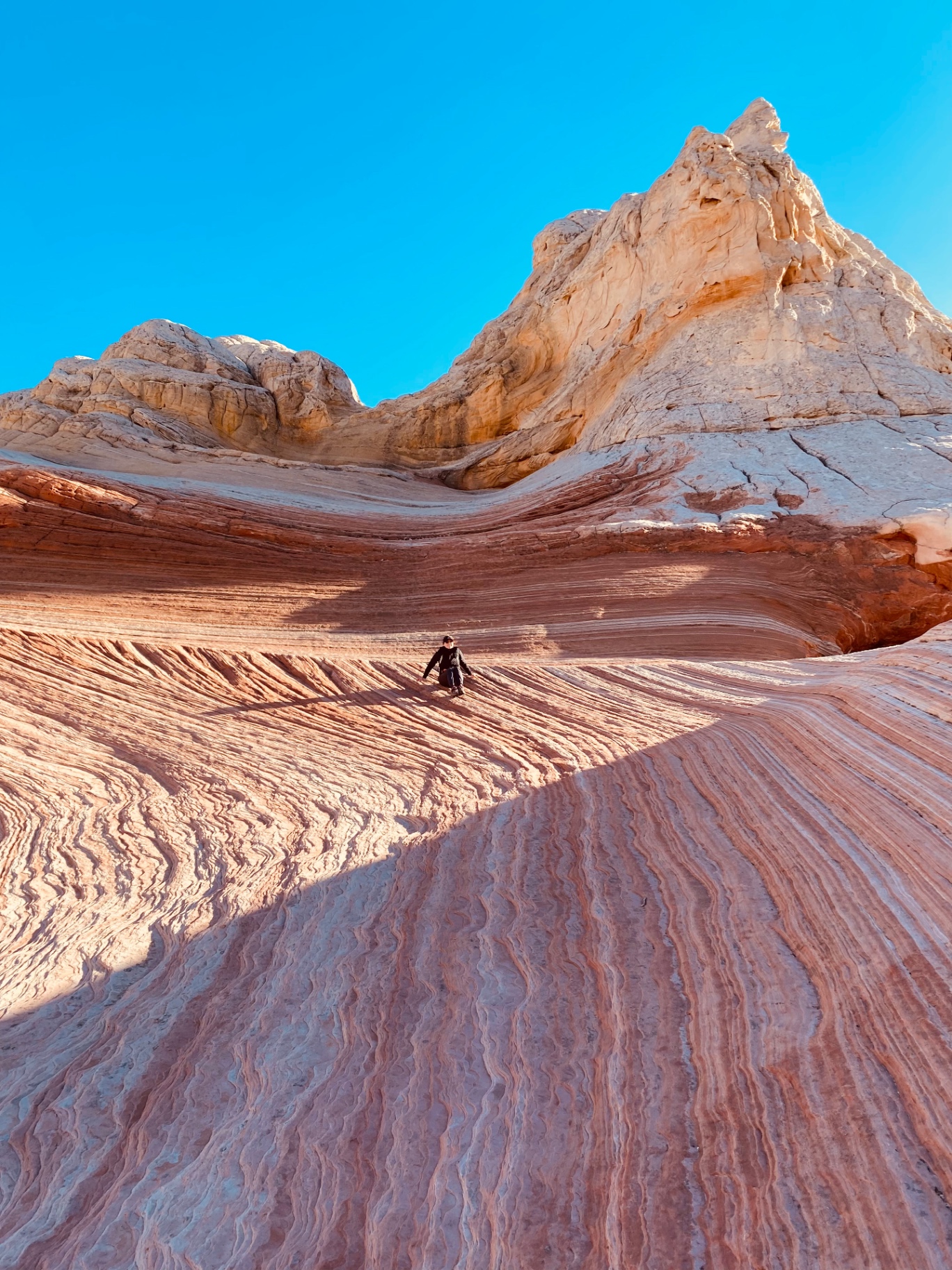 A lone figure in dark clothing on a sweeping slope of rippled sandstone, its cream, salmon and terracotta bands curving up toward pale buttes under a cloudless deep blue sky, a long shadow crossing the stone